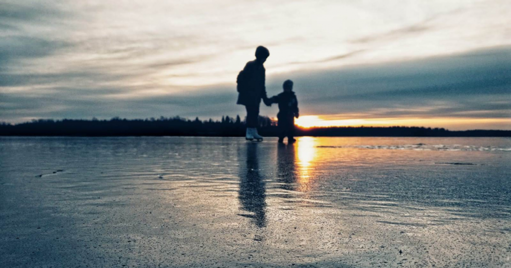 Kaksi lasta luistelee jäällä auringonlaskua vasten.